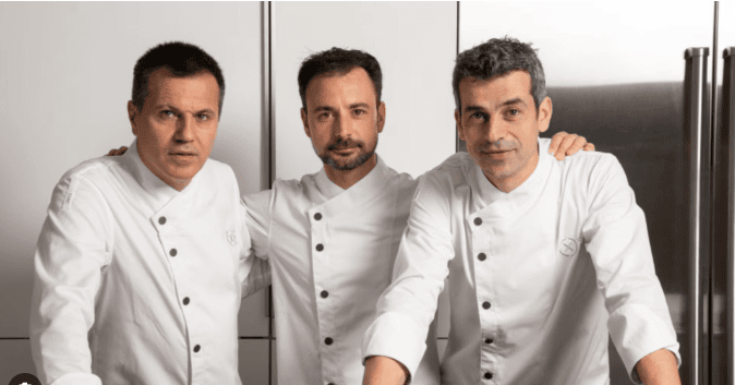 Three chefs in white uniforms stand together in a kitchen, two with their arms around each other. They are looking at the camera with neutral expressions. The background features metal cabinets and a refrigerator.